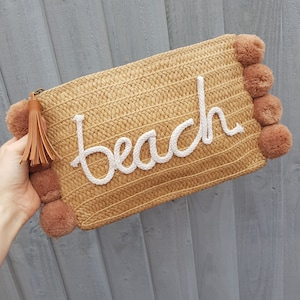 May include: A straw clutch bag with a white "beach" embroidered on the front. The bag has brown pom-poms on the sides and a brown tassel.