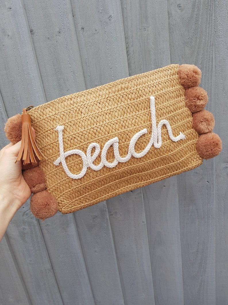 May include: A straw clutch bag with a white "beach" embroidered on the front. The bag has brown pom-poms on the sides and a brown tassel.