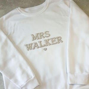 May include: White sweatshirt with the text "MRS WALKER" in silver rhinestones. A small rhinestone heart is below the name. The sweatshirt has a crew neck and ribbed cuffs.