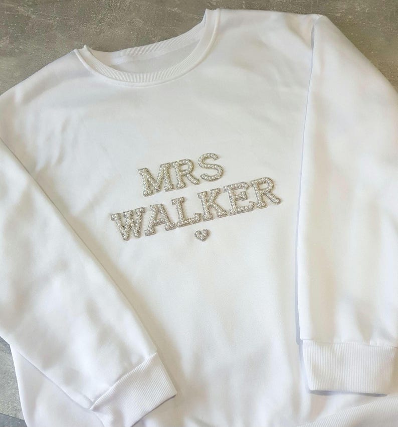 May include: White sweatshirt with the text "MRS WALKER" in silver rhinestones. A small rhinestone heart is below the name. The sweatshirt has a crew neck and ribbed cuffs.