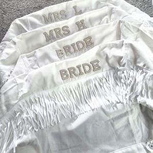May include: Four white denim jackets, each with rhinestone lettering. Two jackets say "BRIDE" and two say "MRS L" and "MRS H". The jackets have fringe detailing across the back.