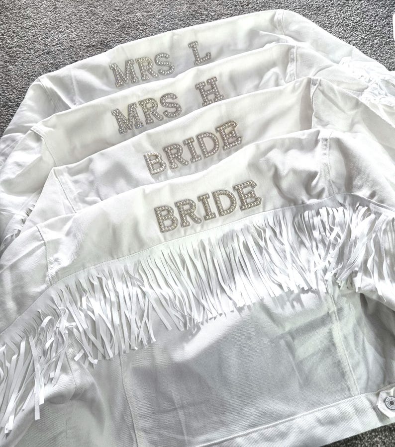 May include: Four white denim jackets, each with rhinestone lettering. Two jackets say "BRIDE" and two say "MRS L" and "MRS H". The jackets have fringe detailing across the back.
