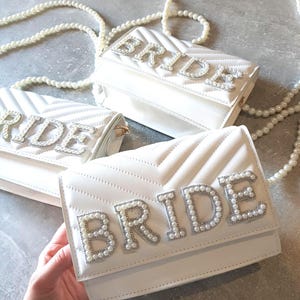 May include: Three white quilted purses with a pearl embellished "BRIDE" design. The purses have a pearl chain strap.