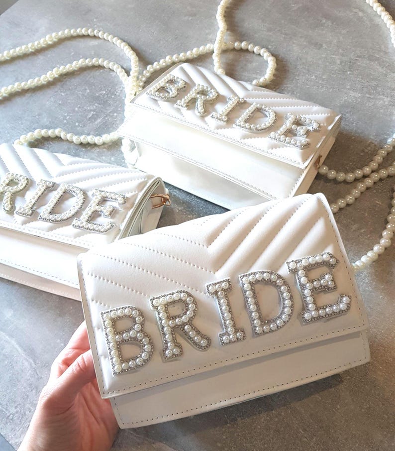 May include: Three white quilted purses with a pearl embellished "BRIDE" design. The purses have a pearl chain strap.