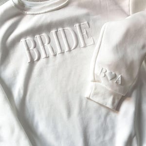 May include: White sweatshirt with the word "BRIDE" embossed across the chest. The sleeve cuff features the initials "B♡A". The crew neck sweatshirt is a bridal themed apparel item.