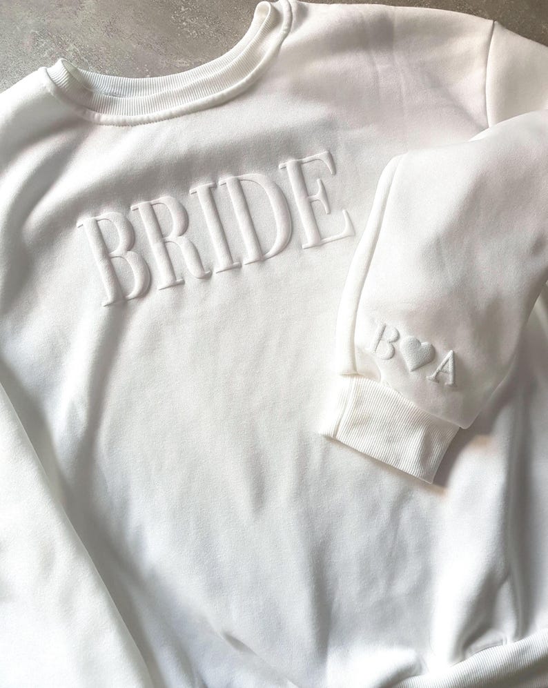 May include: White sweatshirt with the word "BRIDE" embossed across the chest. The sleeve cuff features the initials "B♡A". The crew neck sweatshirt is a bridal themed apparel item.