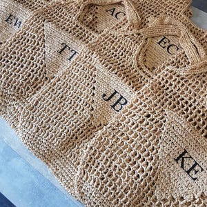May include: A collection of tan crochet tote bags featuring an open-weave design and two handles. Each bag displays a triangular patch with black embroidered initials, including EW, CE, AC, TT, EC, JB, and KE, arranged in a layered group.