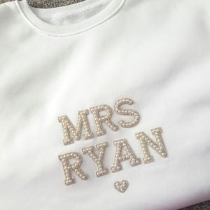 May include: White crewneck sweatshirt with the text "MRS RYAN" in silver and pearl embellishments. A small heart is below the name. The sweatshirt is a soft, casual garment.