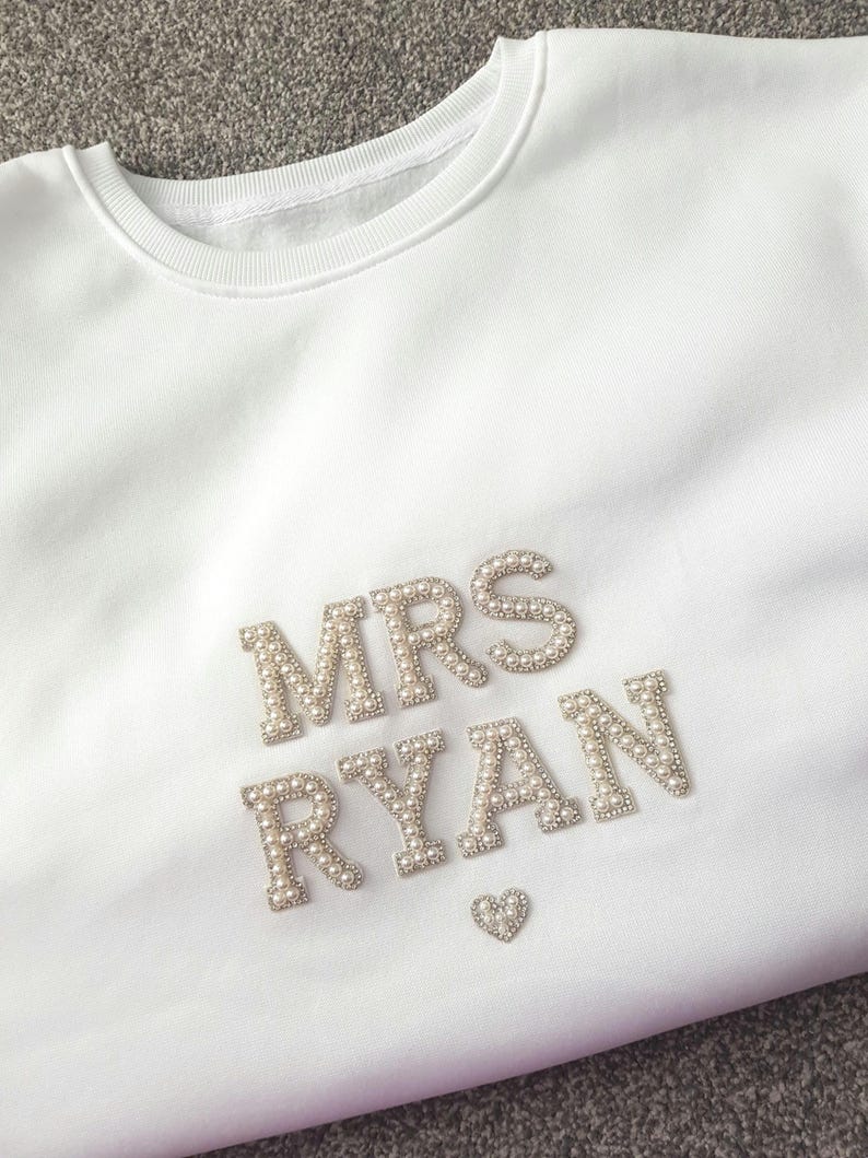 May include: White crewneck sweatshirt with the text "MRS RYAN" in silver and pearl embellishments. A small heart is below the name. The sweatshirt is a soft, casual garment.