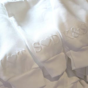 May include: Close-up of three white sweatshirts with embroidered initials and heart designs. The initials are in a simple, elegant font. The sweatshirts are folded, showing the cuffs with the embroidered details.