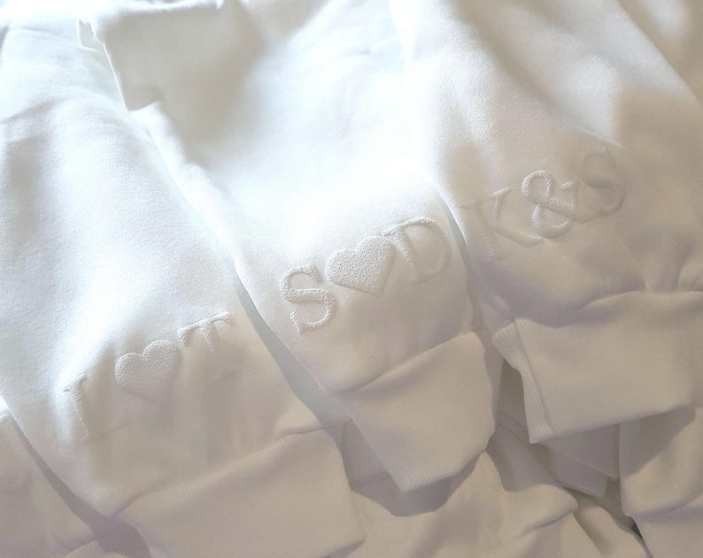 May include: Close-up of three white sweatshirts with embroidered initials and heart designs. The initials are in a simple, elegant font. The sweatshirts are folded, showing the cuffs with the embroidered details.