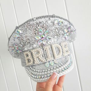 Op de afbeelding: Zilveren paillettenpet met het woord "BRIDE" gespeld in parelachtige kralen. De pet is versierd met iriserende strass-steentjes en parels. De pet is een bruidsaccessoire.