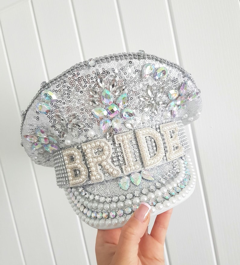 Op de afbeelding: Zilveren paillettenpet met het woord "BRIDE" gespeld in parelachtige kralen. De pet is versierd met iriserende strass-steentjes en parels. De pet is een bruidsaccessoire.