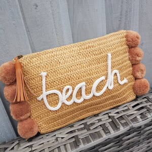 May include: A straw clutch bag with a brown tassel and pom-poms. The bag has the word "Beach" embroidered on it in white.