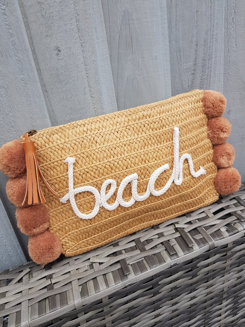 May include: A straw clutch bag with a brown tassel and pom-poms. The bag has the word "Beach" embroidered on it in white.