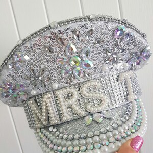 Op de afbeelding: Een zilveren pailletten hoed met het woord "MRS" uitgespeld in grote, witte parels. De hoed is versierd met iriserende strass en parels. De rand is afgezet met parels.