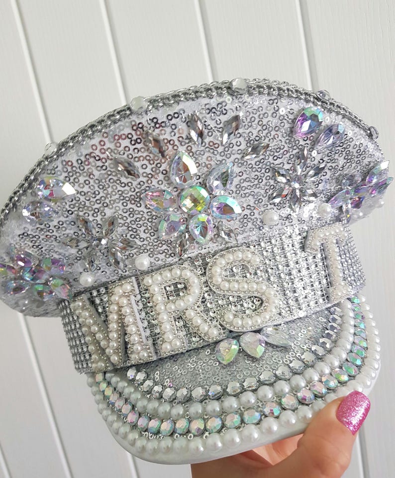 Op de afbeelding: Een zilveren pailletten hoed met het woord "MRS" uitgespeld in grote, witte parels. De hoed is versierd met iriserende strass en parels. De rand is afgezet met parels.