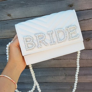 May include: White quilted clutch bag with a pearl embellished "BRIDE" design and a pearl chain strap.