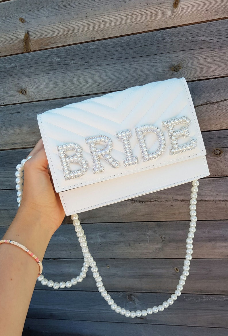 May include: White quilted clutch bag with a pearl embellished "BRIDE" design and a pearl chain strap.