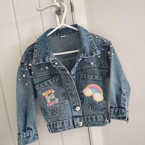 May include: A blue denim jacket with pearl embellishments. The jacket has a rainbow and cloud patch on one pocket and a colourful letter "B" patch on the other pocket.
