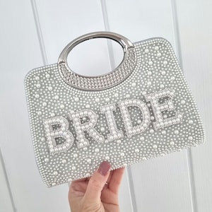 May include: Silver clutch bag covered in pearls, with a silver handle. The word "BRIDE" is spelled out in large pearl letters. A stylish accessory for a wedding or bridal event.