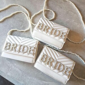 May include: Three white quilted purses with pearl straps. Each purse has the word "BRIDE" spelled out in rhinestones.