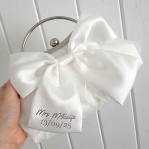 May include: A white satin clutch bag with a silver clasp and a bow. The bag is personalized with the text 'Mrs. Metcalfe 13/09/25'.