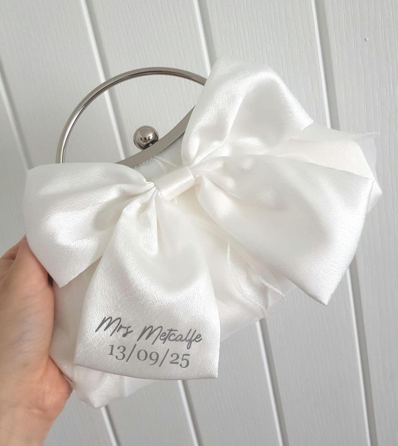 May include: A white satin clutch bag with a silver clasp and a bow. The bag is personalized with the text 'Mrs. Metcalfe 13/09/25'.