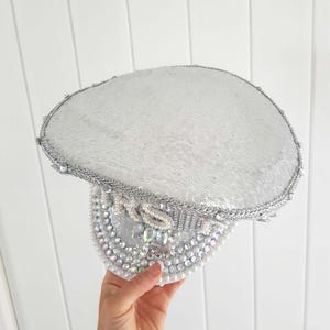 Op de afbeelding: Een zilveren hoed met pailletten, een afgeronde bovenkant en een gebogen klep. De klep is rijkelijk versierd met parels, pailletten en iriserende edelstenen. De hoed is afgezet met zilveren vlechtwerk en heeft het woord "MRS" in parels.