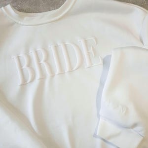 May include: A white sweatshirt with the word "BRIDE" in raised, textured lettering across the chest. The crew neck and cuffs are visible. The garment is made of a soft, likely cotton, material.