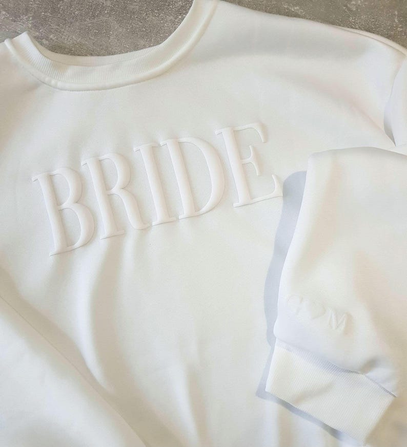 May include: A white sweatshirt with the word "BRIDE" in raised, textured lettering across the chest. The crew neck and cuffs are visible. The garment is made of a soft, likely cotton, material.