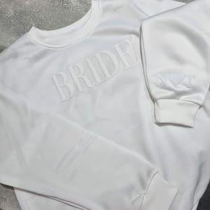 May include: A white sweatshirt with the word "BRIDE" embossed across the chest. The sleeve cuff features the word "NOT" and a heart symbol. The sweatshirt has a crew neck and ribbed cuffs.