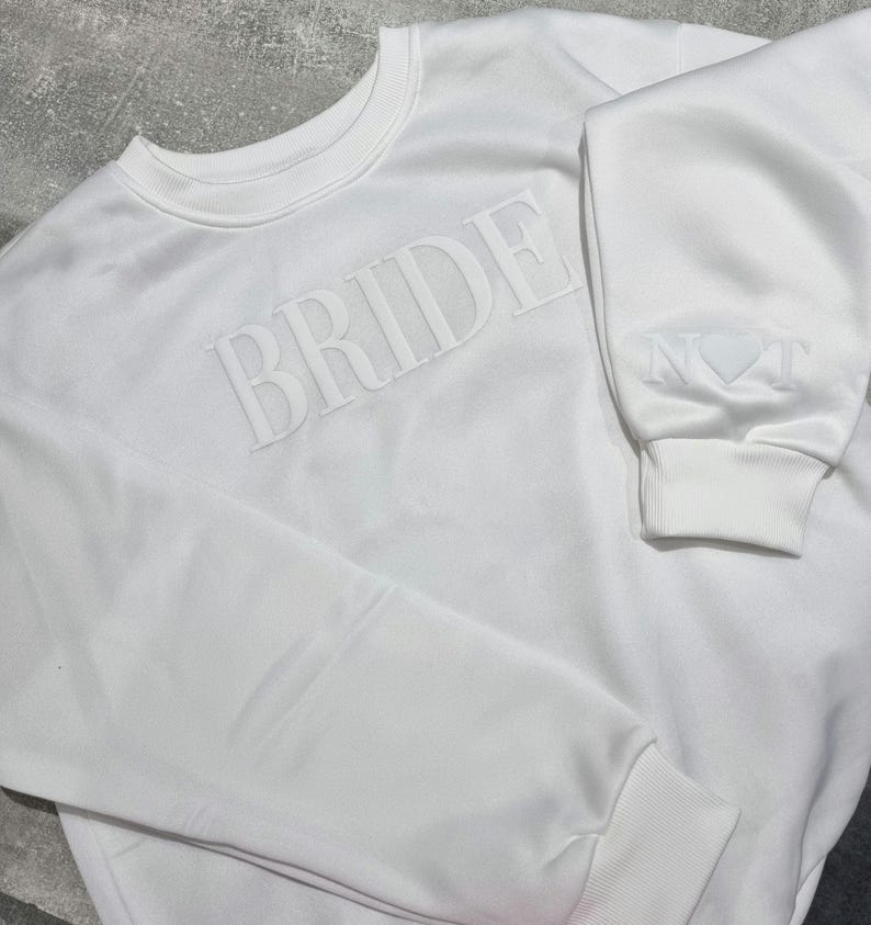 May include: A white sweatshirt with the word "BRIDE" embossed across the chest. The sleeve cuff features the word "NOT" and a heart symbol. The sweatshirt has a crew neck and ribbed cuffs.