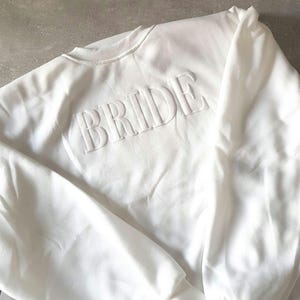 May include: A white crewneck sweatshirt with the word "BRIDE" embossed across the chest. The sweatshirt is made of a soft material and has a ribbed collar and cuffs. The overall style is simple and elegant, perfect for bridal wear.