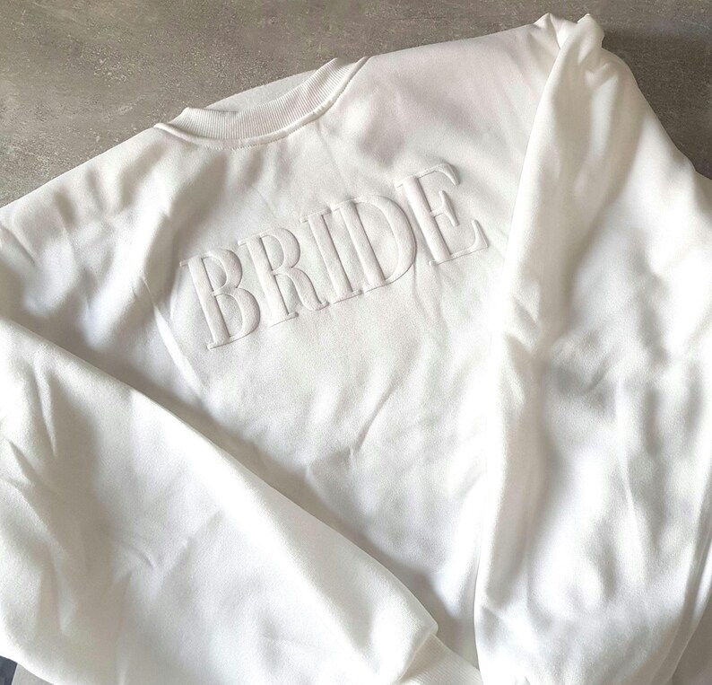 May include: A white crewneck sweatshirt with the word "BRIDE" embossed across the chest. The sweatshirt is made of a soft material and has a ribbed collar and cuffs. The overall style is simple and elegant, perfect for bridal wear.