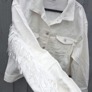 May include: Off-white denim jacket with fringe detail on the sleeves. The jacket has a classic collar, button-down front, and two chest pockets. The XL size tag is visible. The jacket is hanging on a black hanger.