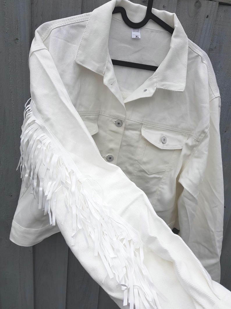 May include: Off-white denim jacket with fringe detail on the sleeves. The jacket has a classic collar, button-down front, and two chest pockets. The XL size tag is visible. The jacket is hanging on a black hanger.