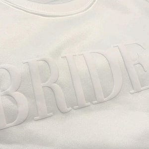 May include: White crew neck sweatshirt with the word "BRIDE" in raised, tonal lettering across the chest. The letters are large and prominent, creating a clean and minimalist design. The sweatshirt is made of a soft, textured fabric.