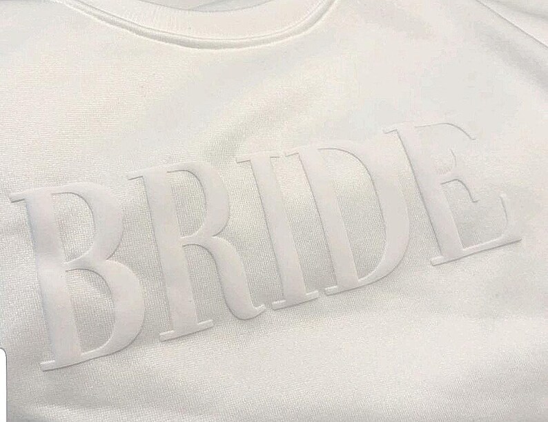 May include: White crew neck sweatshirt with the word "BRIDE" in raised, tonal lettering across the chest. The letters are large and prominent, creating a clean and minimalist design. The sweatshirt is made of a soft, textured fabric.