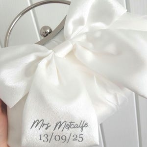 May include: White satin clutch bag with a large bow and a silver clasp. The bag is personalized with the text "Mrs. Metcalfe 13/09/25".