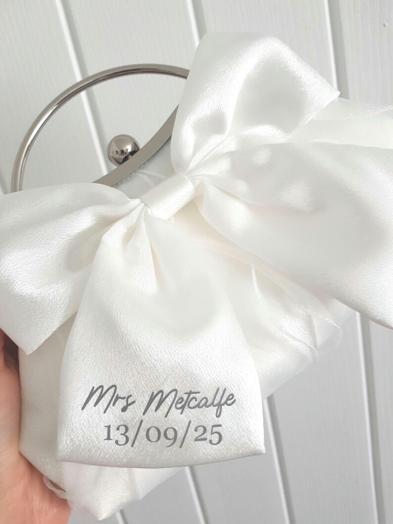 May include: White satin clutch bag with a large bow and a silver clasp. The bag is personalized with the text "Mrs. Metcalfe 13/09/25".