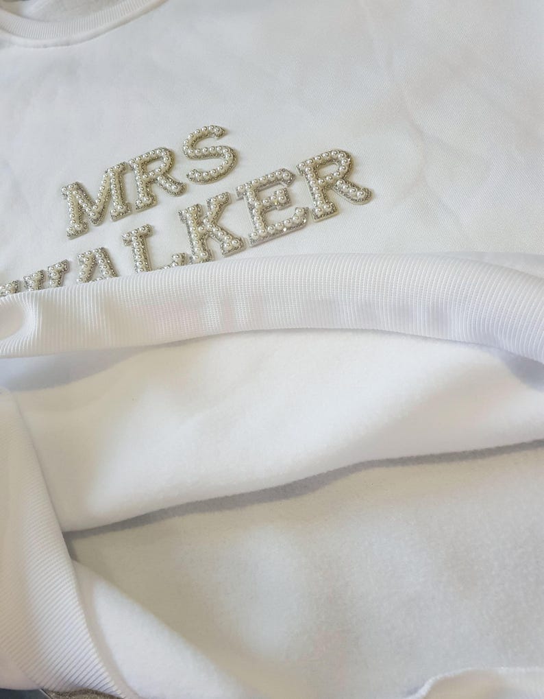 May include: White sweatshirt with the text "MRS WALKER" in pearl-like embellishments. The sweatshirt has a soft, fleecy interior and a ribbed neckline. The design is centered on the upper chest.
