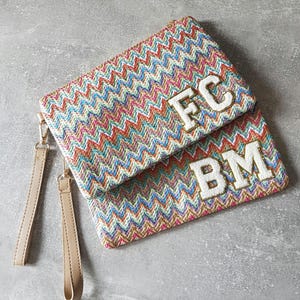 May include: A colourful woven clutch bag with two straps. The bag has the initials "FC" and "BM" embroidered on the front in white.