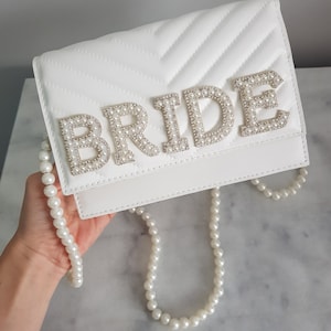 May include: White quilted clutch bag with a pearl embellished "BRIDE" design. The bag has a pearl chain strap.
