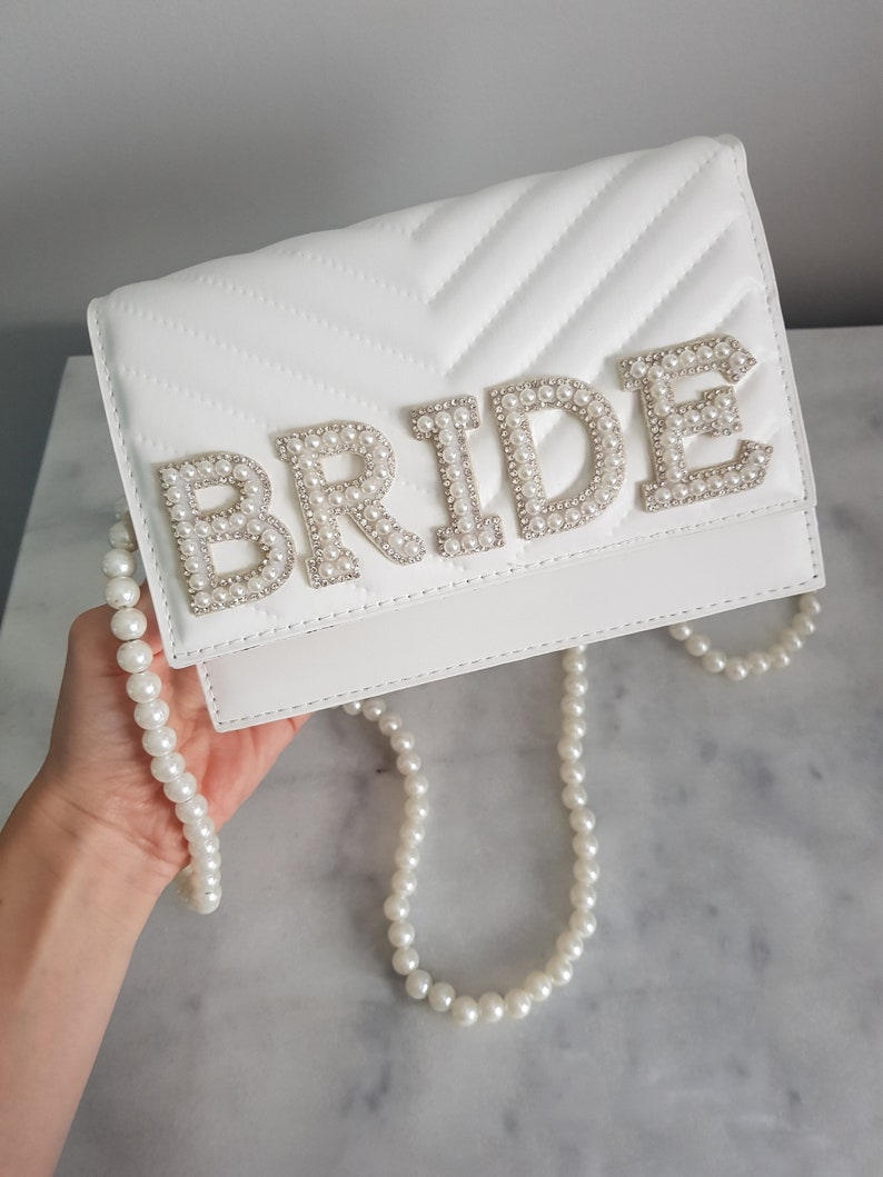 May include: White quilted clutch bag with a pearl embellished "BRIDE" design. The bag has a pearl chain strap.