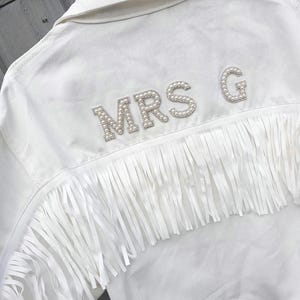 May include: White denim jacket with fringe detail and the text "MRS G" spelled out in pearl-like embellishments. The jacket has a classic collar and a row of fringe across the back.