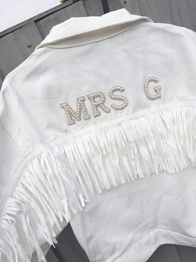 May include: White denim jacket with fringe detail and the text "MRS G" spelled out in pearl-like embellishments. The jacket has a classic collar and a row of fringe across the back.