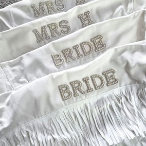 May include: White denim jackets with rhinestone lettering spelling out "MRS L", "MRS H", and "BRIDE". One jacket features fringe along the bottom. Perfect for a bridal party.