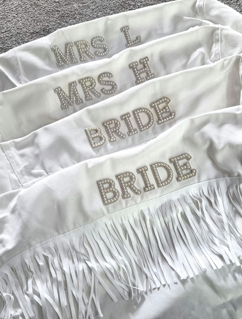 May include: White denim jackets with rhinestone lettering spelling out "MRS L", "MRS H", and "BRIDE". One jacket features fringe along the bottom. Perfect for a bridal party.