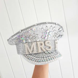 Op de afbeelding: Zilveren pailletten hoed met het woord "MRS" in parelachtige kralen. De hoed is versierd met iriserende strass-steentjes en parels. De hoed heeft een klep en is waarschijnlijk een bruidsaccessoire.